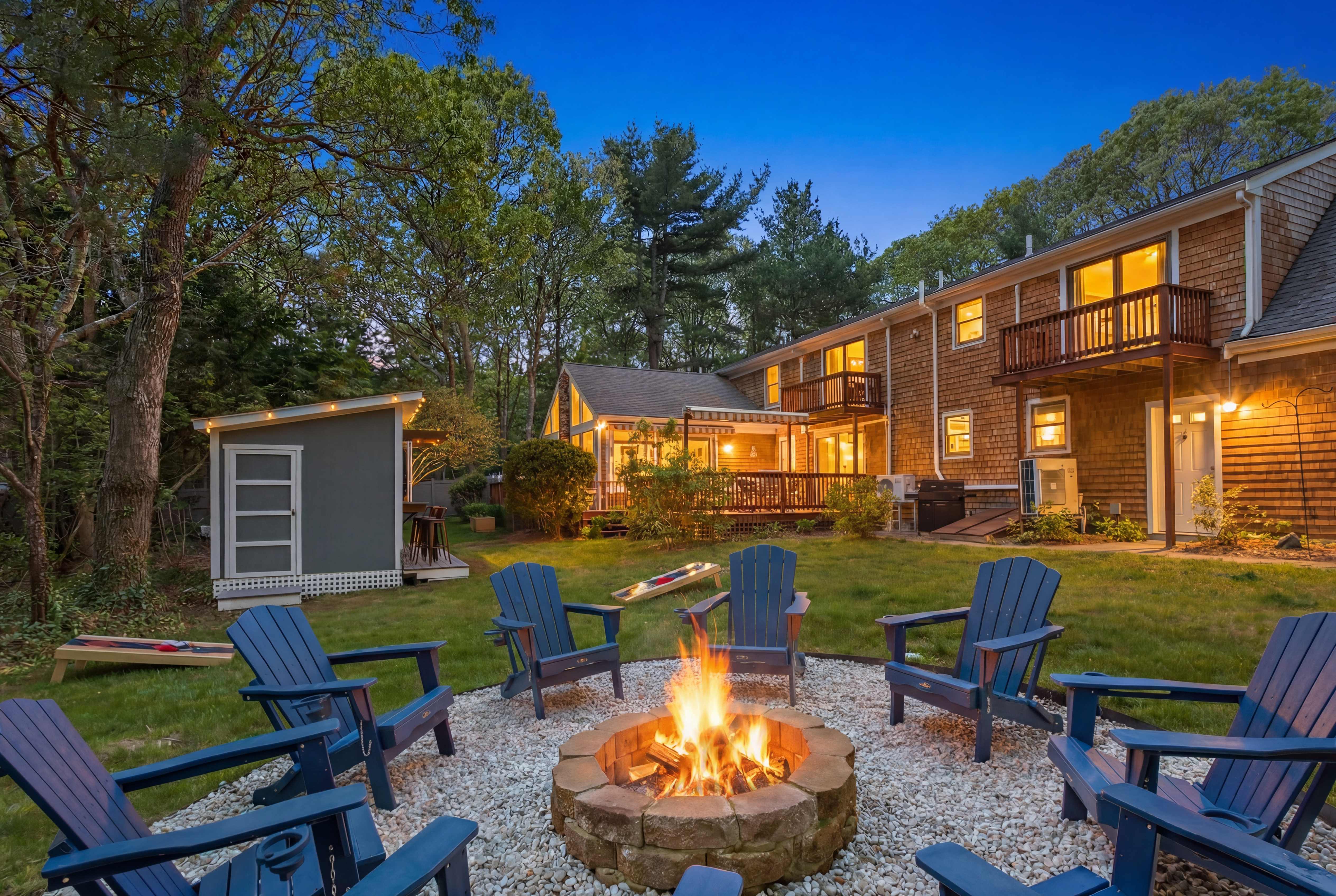 Evening view of the lit fire pit and Adirondack chair circle with cornhole boards on the lawn and every window of the house glowing at dusk
