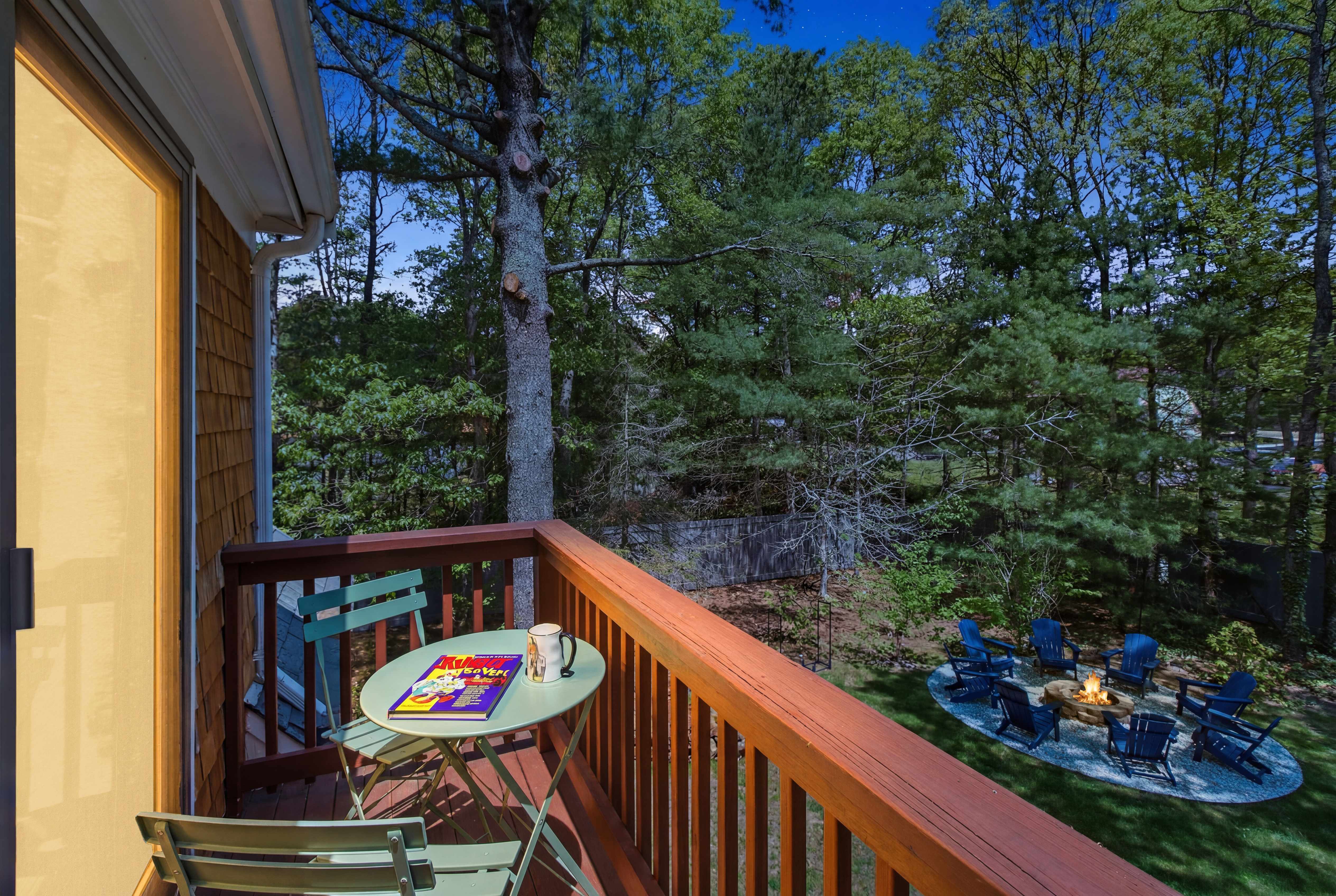 Balcony bistro table with a book and mug at dusk, framing the illuminated fire pit and Adirondack chairs in the wooded backyard below