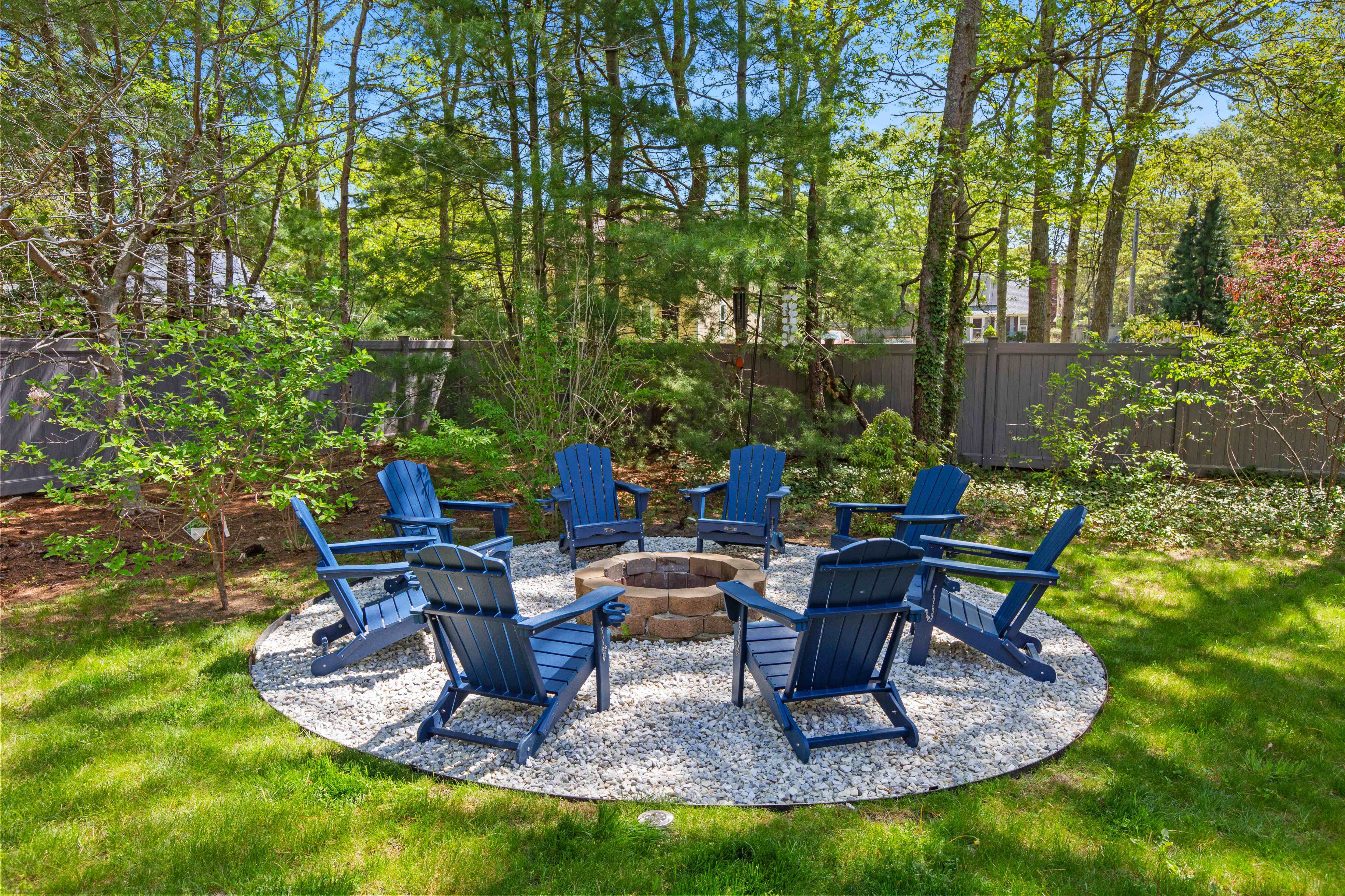Circle of eight navy Adirondack chairs around a stone fire pit set on a bed of white gravel, ringed by tall trees
