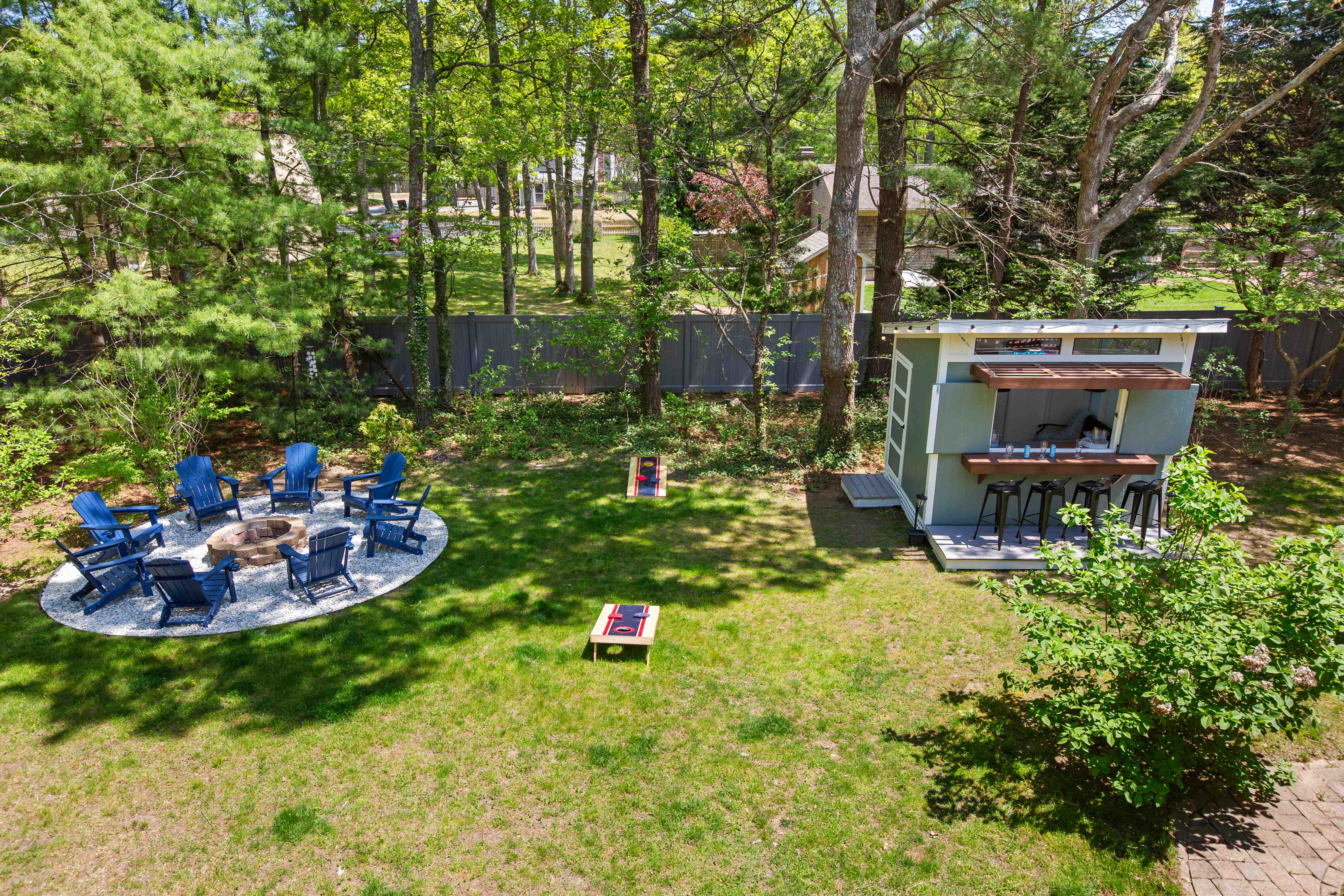Elevated view of the fenced backyard with the fire-pit lounge, two cornhole boards on the lawn, and the bar shed with stools