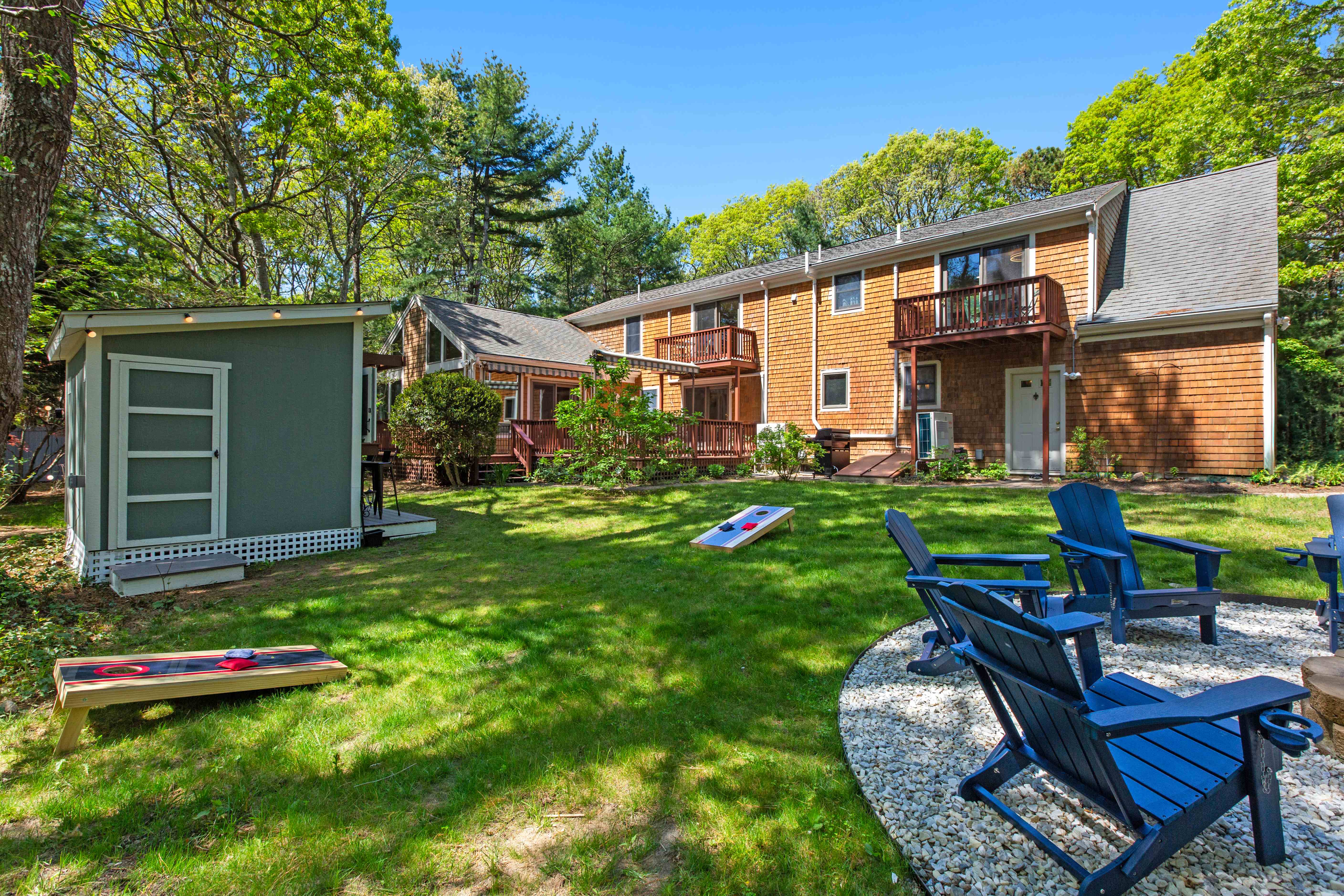 Ground-level view of the cedar-shingled home with its decks and balconies, the green bar shed, cornhole boards, and gravel fire-pit seating