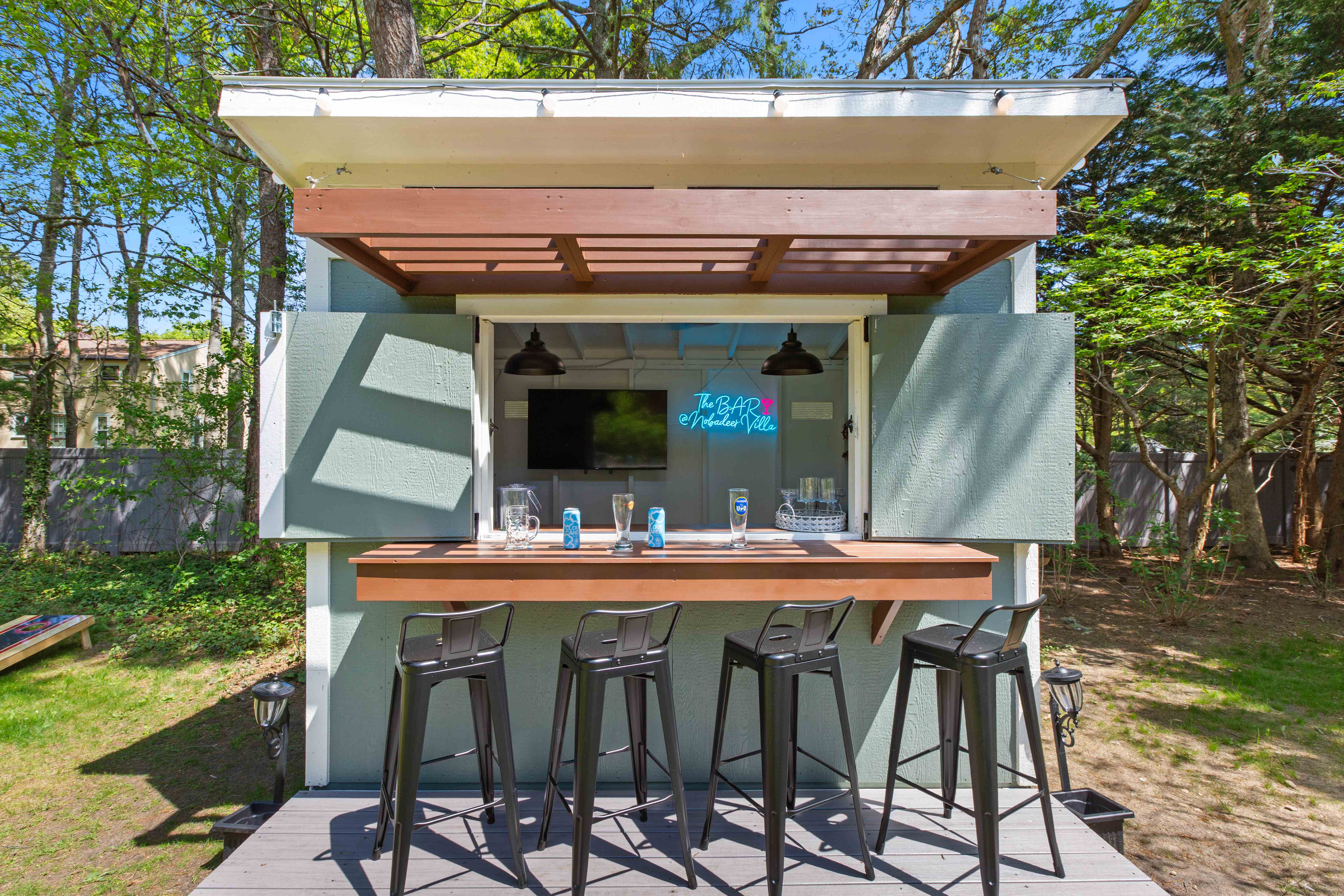 Sage-green backyard bar shed with folding shutters open, a mounted TV, neon sign, and four stools at the wood counter