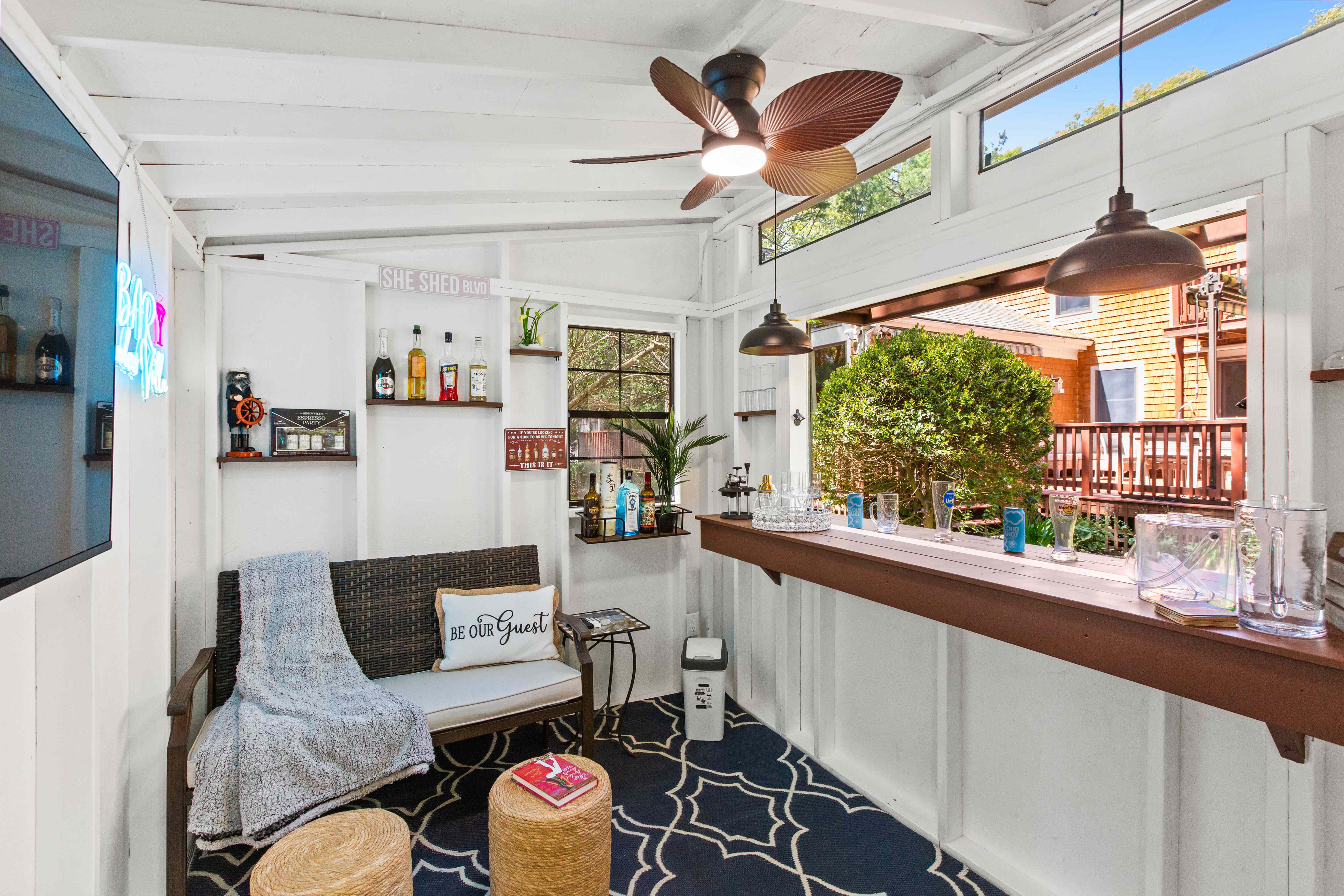 Bar shed interior lounge with a wicker loveseat, liquor shelves, palm-leaf ceiling fan, and pass-through window