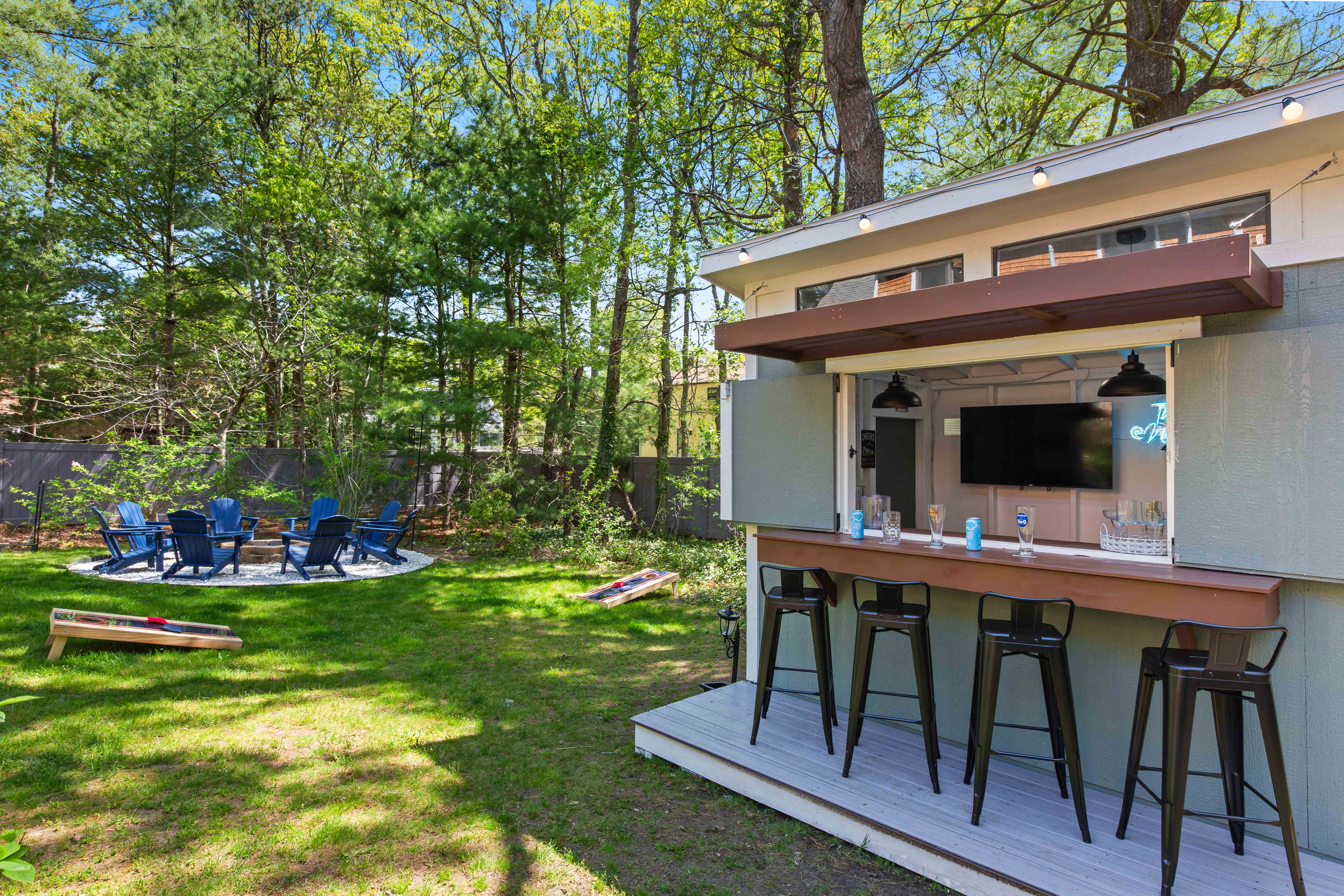 Bar shed and backyard fire-pit circle with blue Adirondack chairs and cornhole boards on the lawn