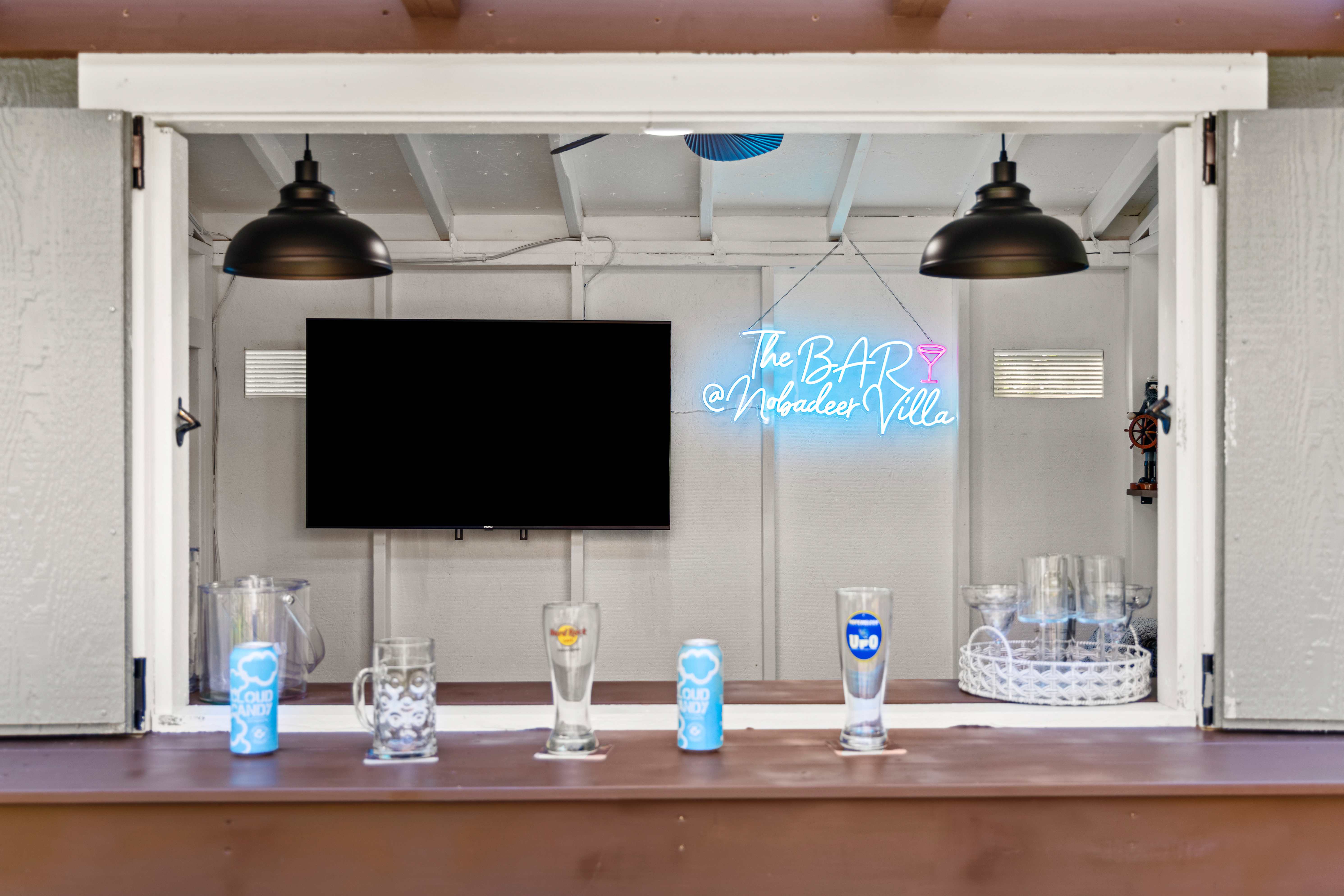 View through the bar window to the mounted TV, neon sign, and pilsner glasses on the counter