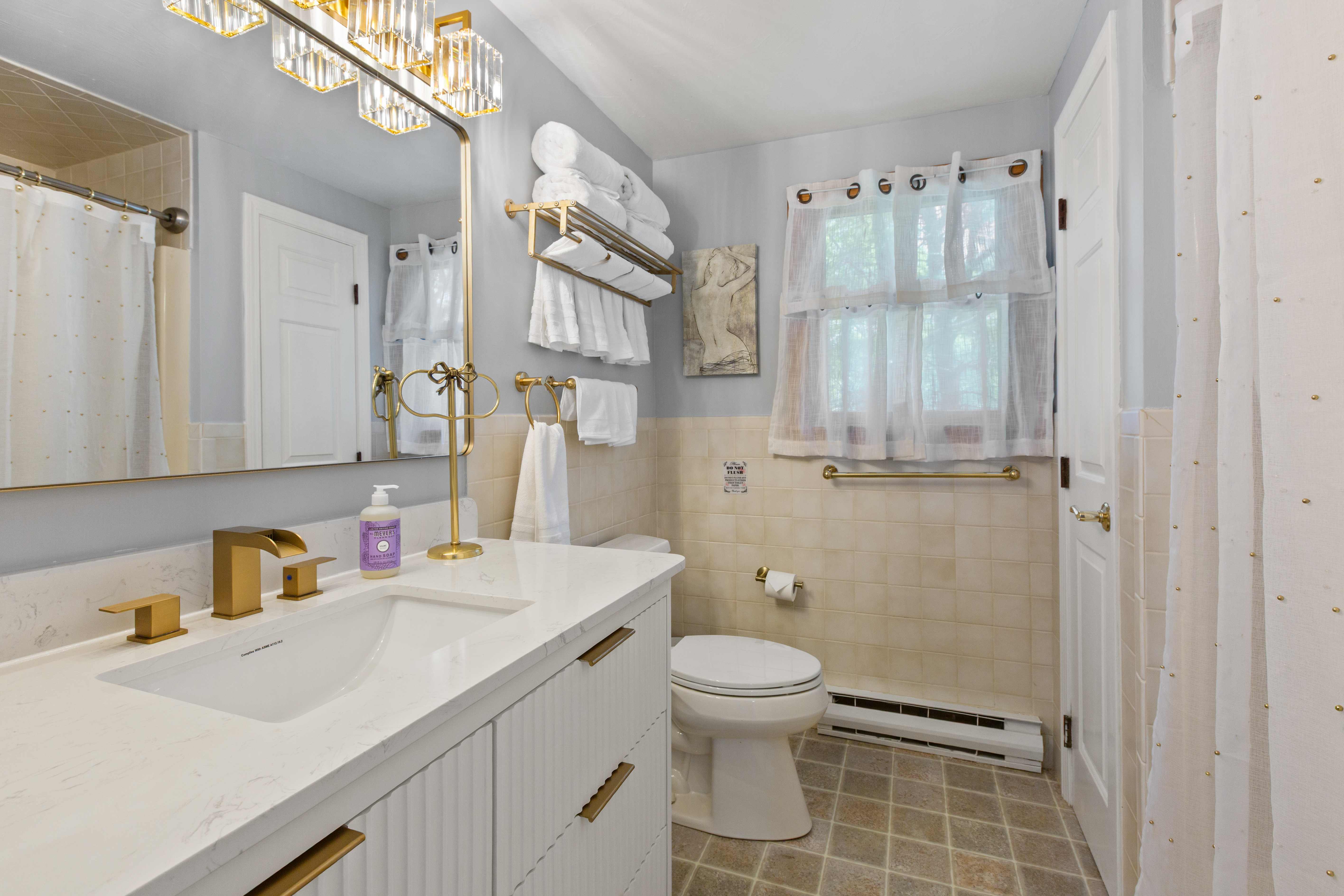 Gray-blue bathroom with a white fluted vanity, gold fixtures, a crystal bar light, and a shower-curtain tub