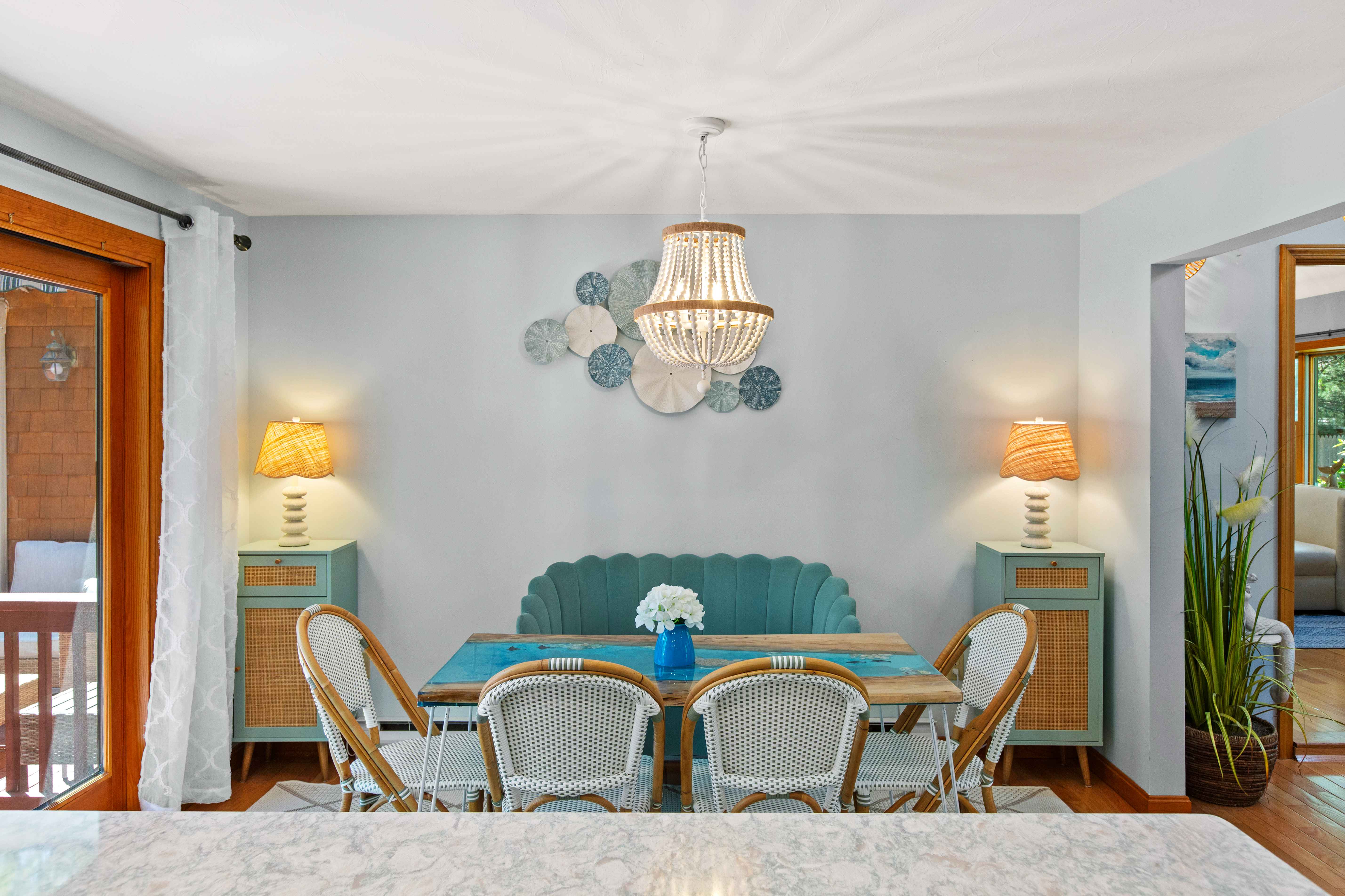 Front view of the breakfast table beneath a white beaded chandelier, framed by teal cane-front cabinets and coastal wall art