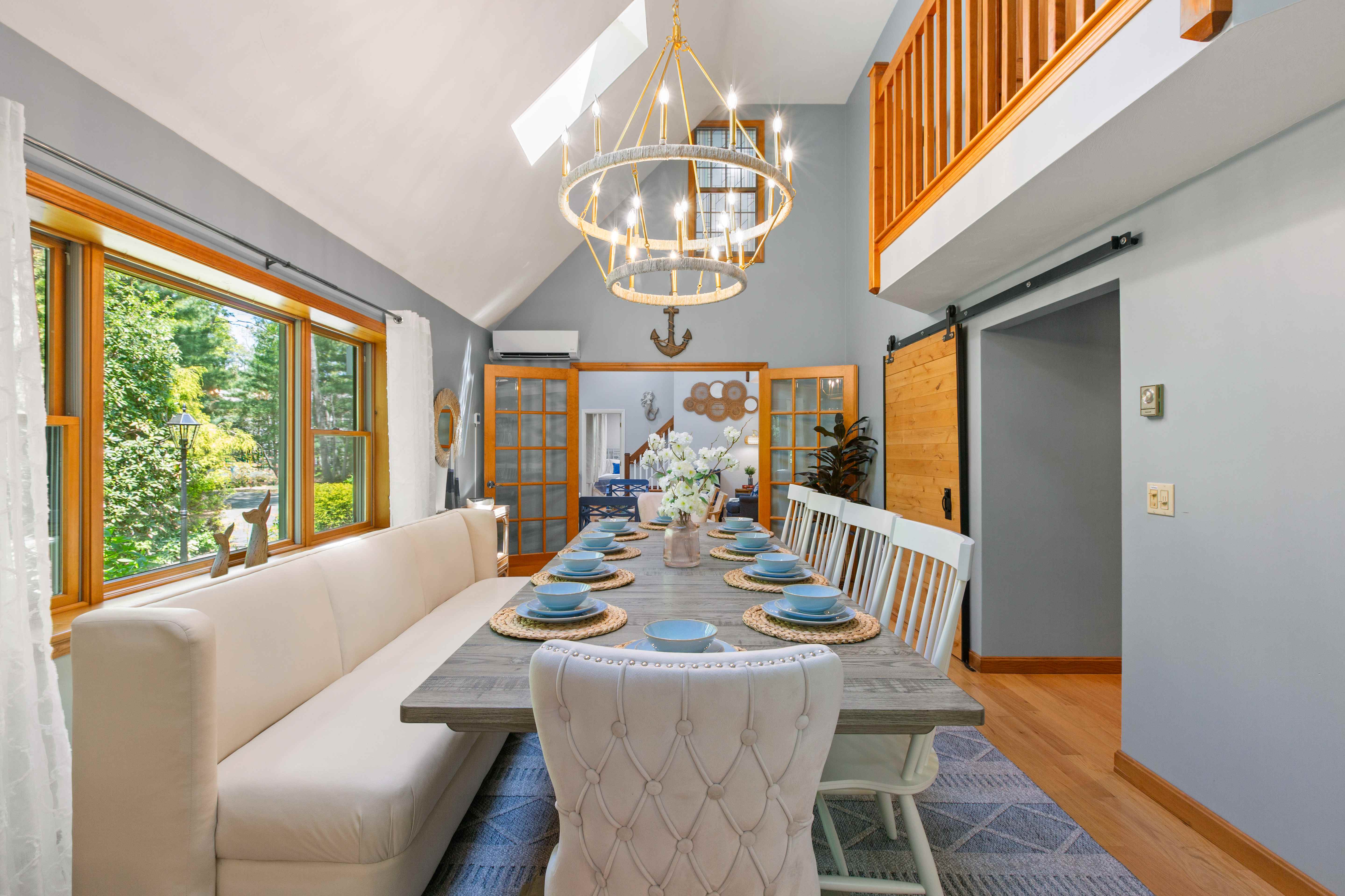 Vaulted dining room with a long weathered-wood table set in blue, a cream banquette under the picture window, and a two-tier rope chandelier