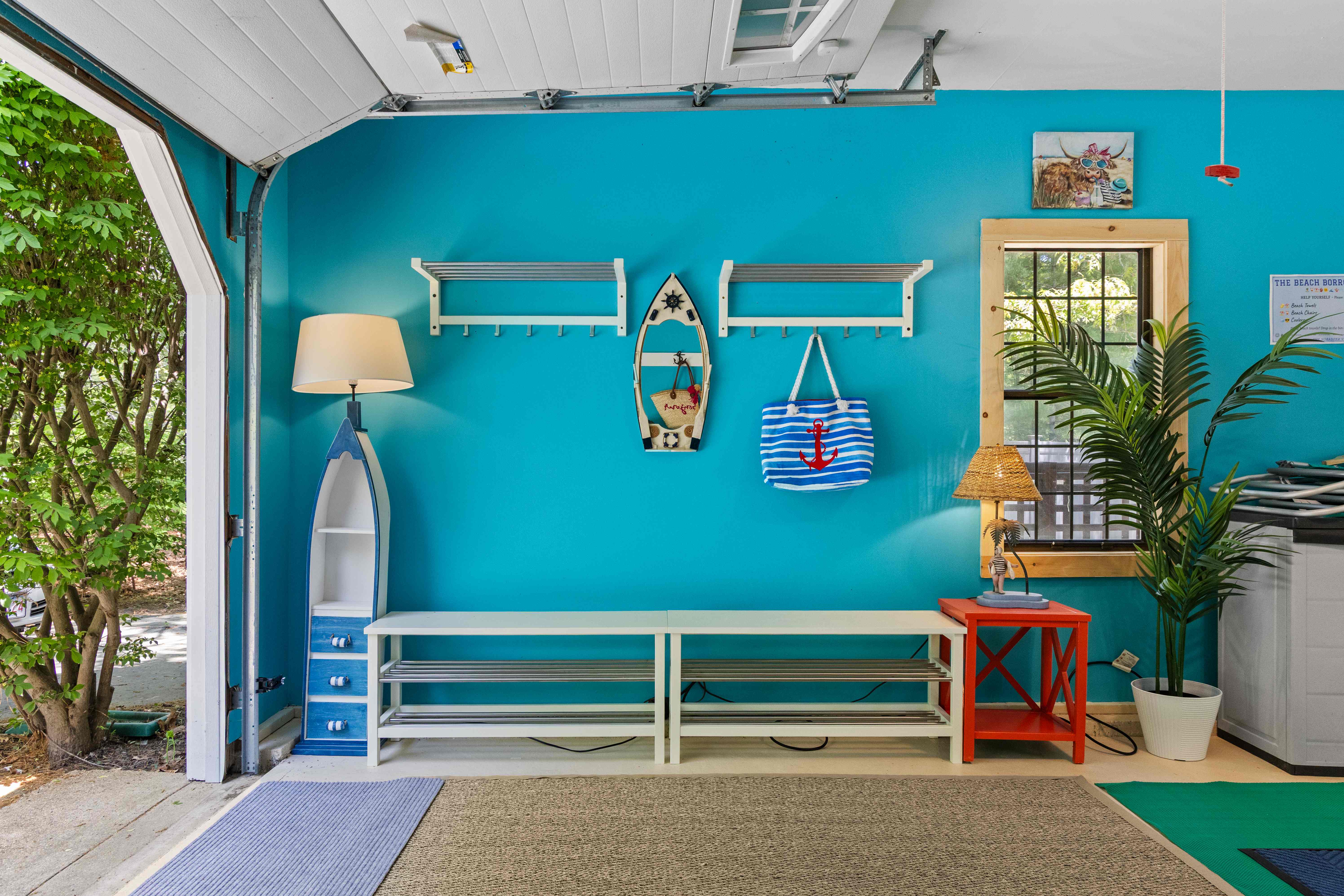 Bright mudroom with white shoe-rack benches, nautical anchor hooks, a boat-shaped shelf, striped beach totes, and the garage door open to greenery