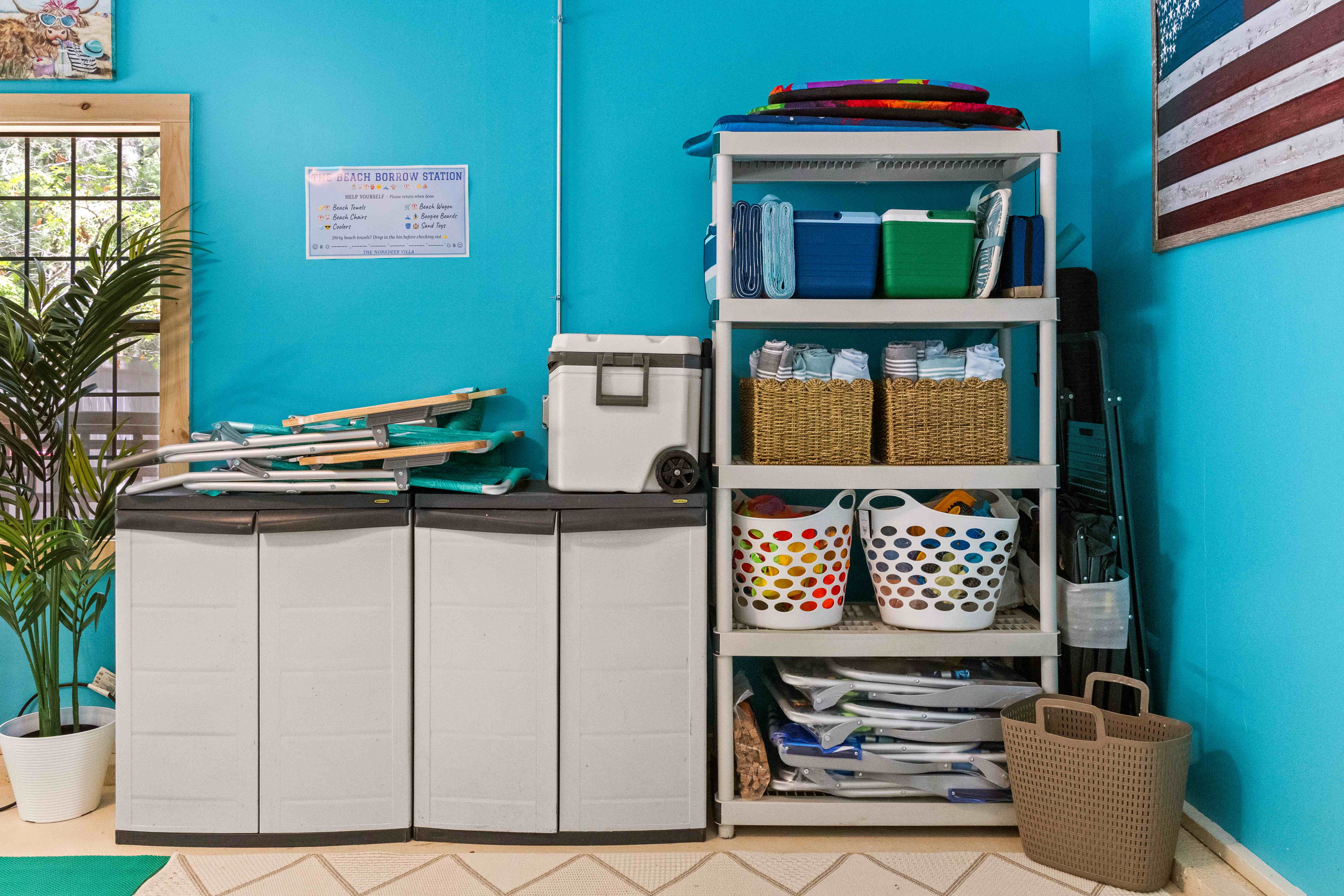 The Beach Borrow Station shelving stocked with coolers, folded towels in woven baskets, beach chairs, and sand toys beside a wheeled Igloo cooler