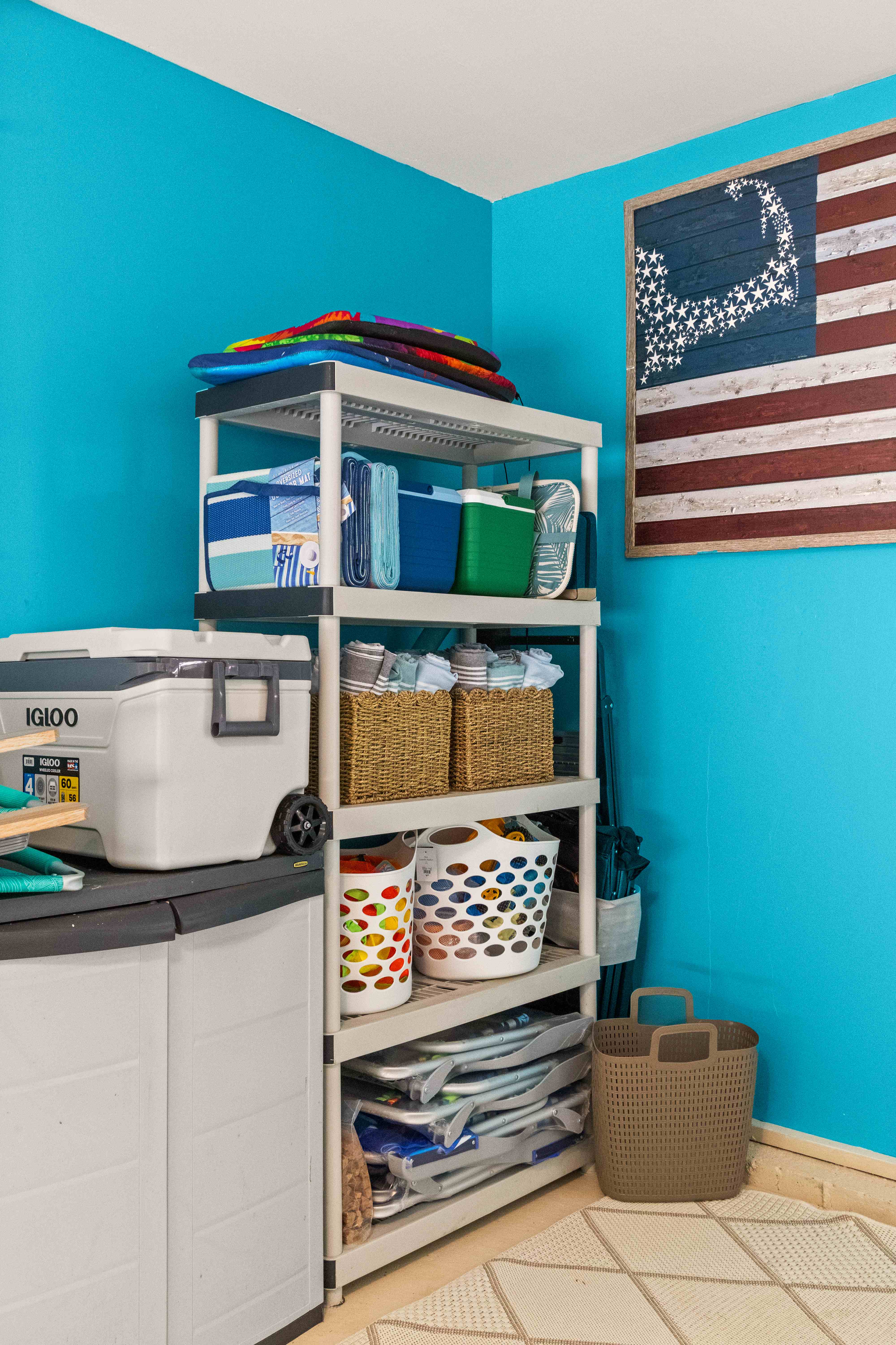 Mudroom corner with a storage shelf of coolers, beach towels in baskets, and folded chairs beneath a framed starfish American flag