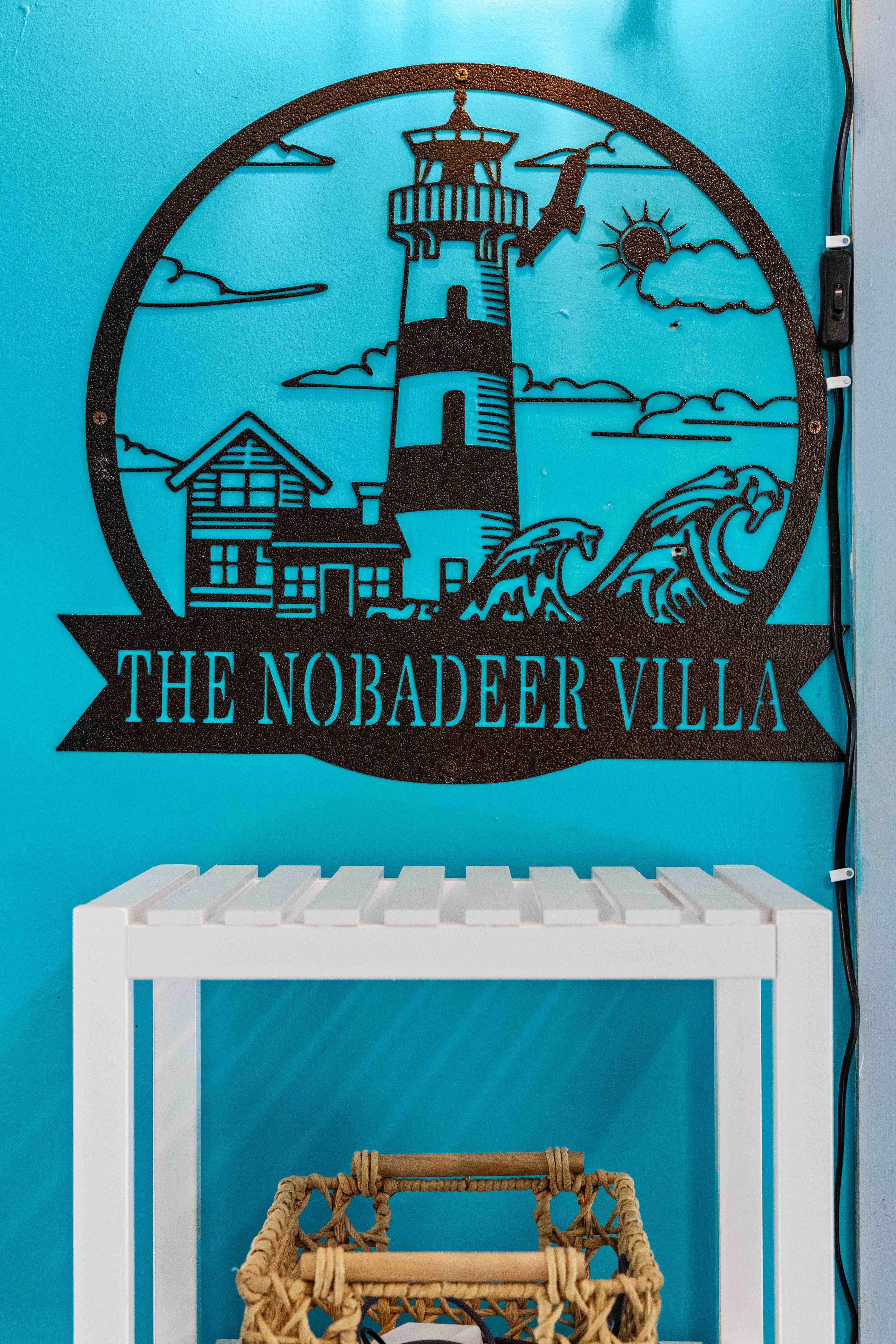 Metal lighthouse sign reading "The Nobadeer Villa" on the turquoise wall above a white slatted console table