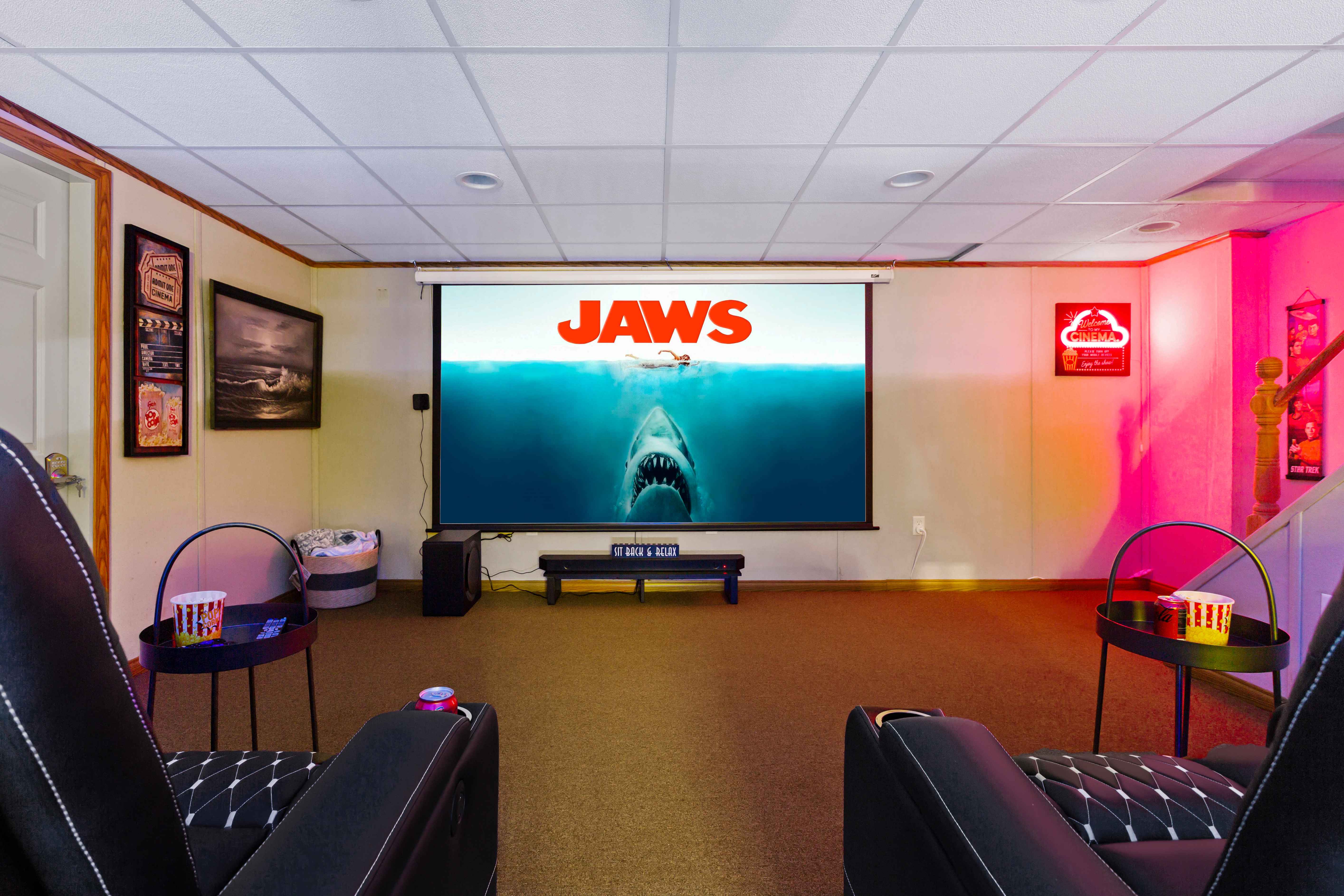Theater projection screen showing the JAWS poster, flanked by recliners, movie posters, and a "Welcome to my Cinema" neon sign