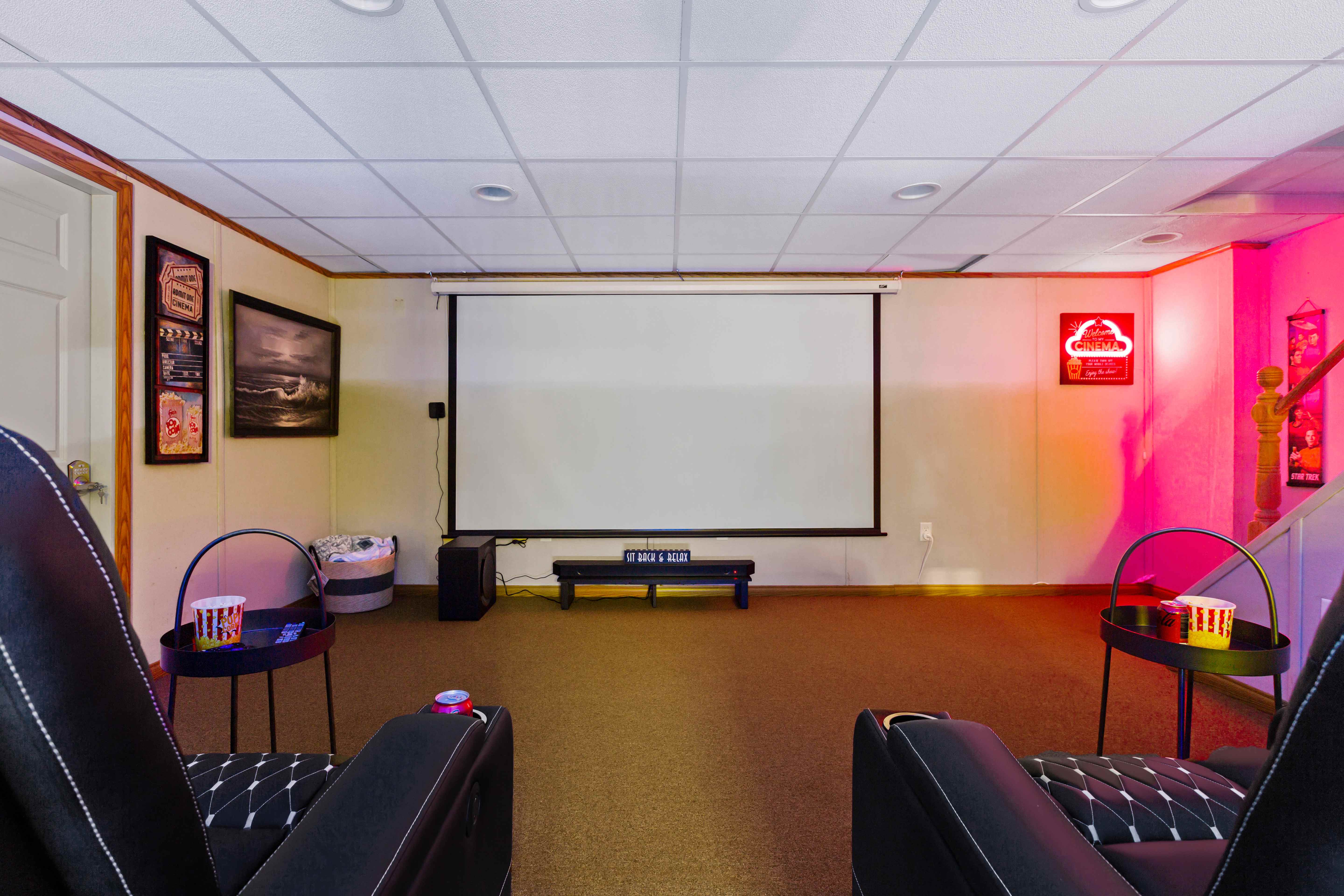 Theater with the projection screen down, plush recliners, side tables set with popcorn and soda, and a cinema neon sign