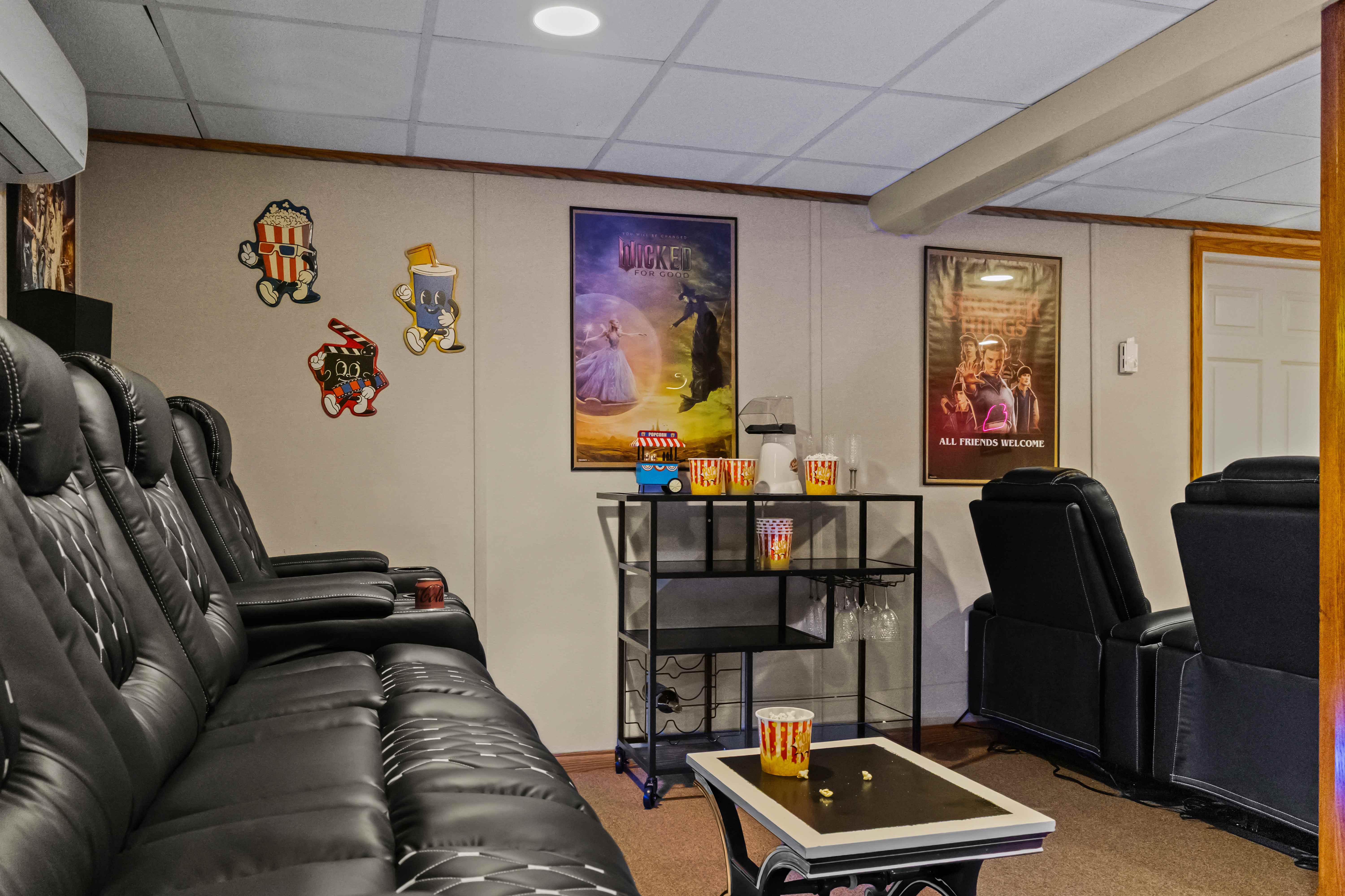 Black leather theater recliners facing a bar cart with a popcorn machine and buckets, beside Wicked and Stranger Things posters